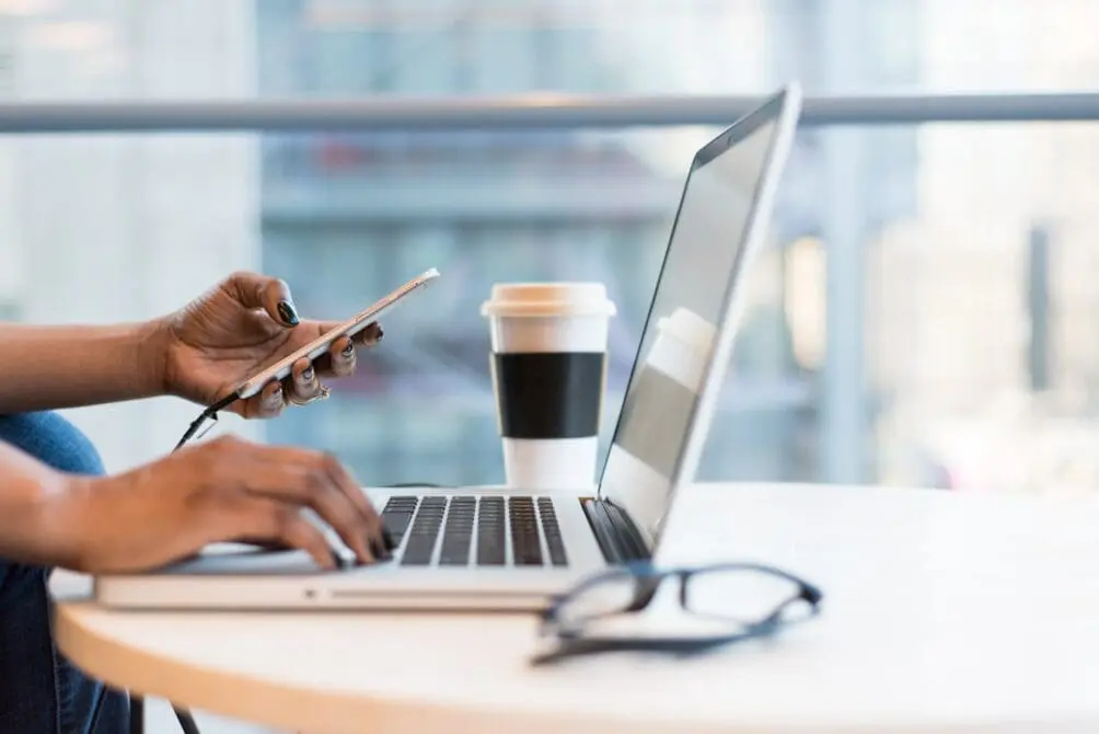 View of a person's hands holding a mobile phone while viewing a laptop