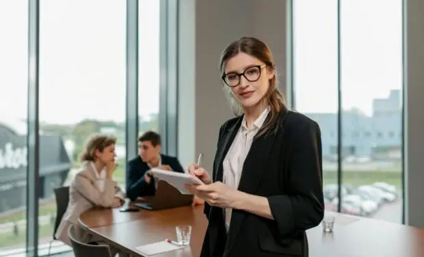 Professional woman in a blazer stands at a conference table, writing in a notebook while colleagues meet in the background. Note: concise, informative ALT text.
