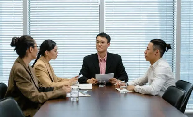 Four professionals in a business meeting around a conference table, reviewing documents as blinds shade the windows behind them.
