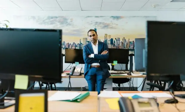 Man in a blue suit stands with arms crossed in a modern open-office, city skyline mural behind him amidst computer monitors and desks, presenting a confident pose.