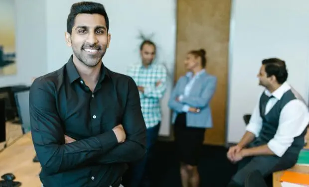 Smiling man in a black shirt with arms crossed at a desk; coworkers chat in the blurred office background.