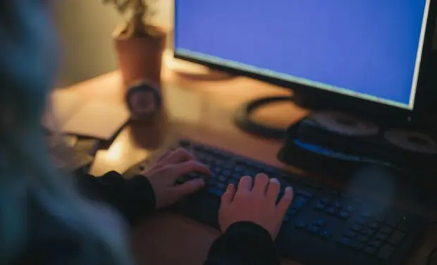 Hands typing on a keyboard at a dimly lit desk with a blue computer monitor in the background.