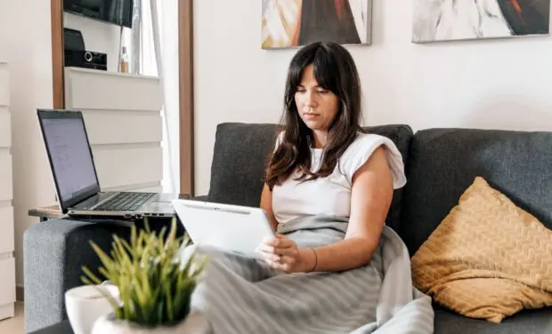 a woman sitting on a couch looking at a tablet with an open laptop nearby