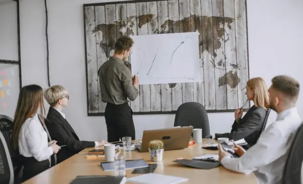 Presenter drawing a graph on a taped sheet attached to a world-map wall as colleagues listen in a conference room.