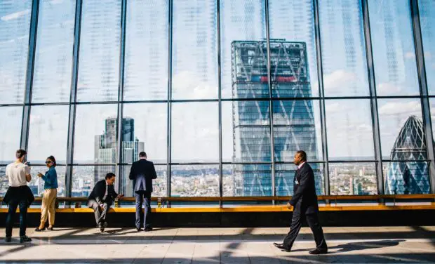 Group of professionals in business attire stand and chat along a glass-walled observation deck overlooking the city skyline.