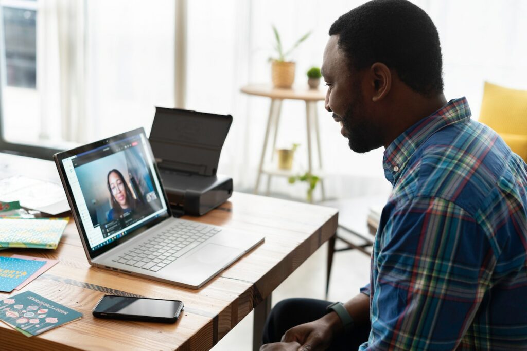 Image of a black man in a plaid shirt man smiling looking at a laptop during a Zoom video call with an image of a woman on the laptop screen. 