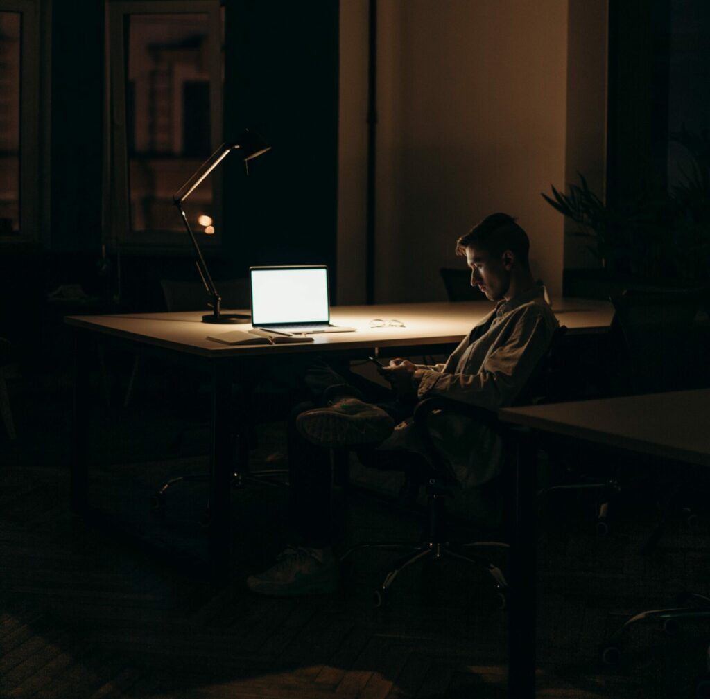 a man sitting alone in an empty office in the dark. One small light illuminates his laptop but the man stares at his phone in his hands tiredly.