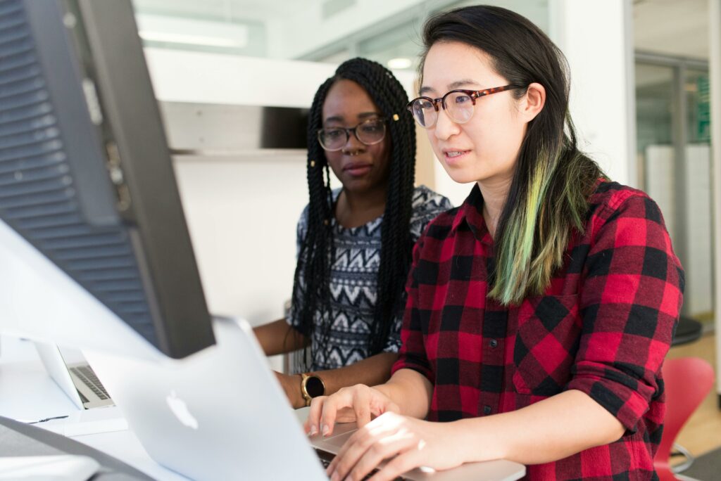 an Asian American woman in a red plaid shirt and an African American woman wearing in a patterned shirt look at a computer monitor together.