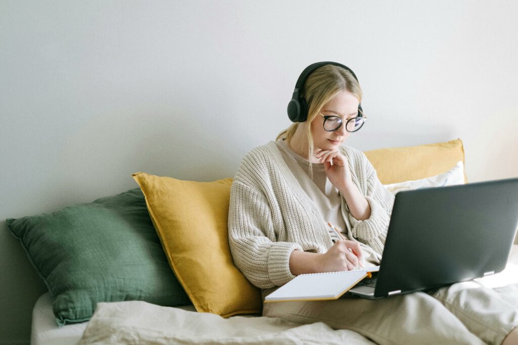woman with glasses sitting on a sofa at home wearing headphones while she writes in a notebook as she has a laptop computer in front of her. 